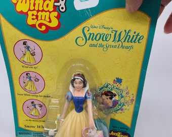 Puede incluir: Un juguete de cuerda de Blancanieves de Disney's Snow White and the Seven Dwarfs. El juguete está vestido con su icónico vestido amarillo y azul y sostiene una cesta de flores. El embalaje es amarillo, verde y blanco con el logotipo de Wind-Ems y el logotipo de Disney.
