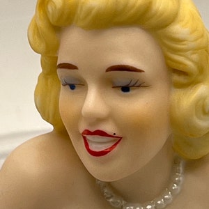 May include: A porcelain figurine of a woman with blonde hair, wearing a pearl necklace and a white dress. The figurine has a smiling expression and blue eyes.