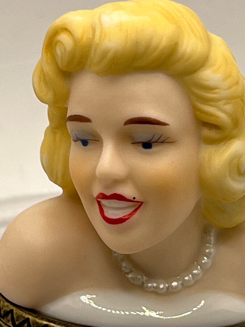 May include: A porcelain figurine of a woman with blonde hair, wearing a pearl necklace and a white dress. The figurine has a smiling expression and blue eyes.