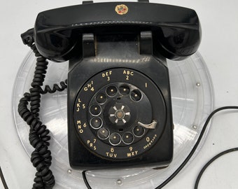 Könnte beinhalten: Ein schwarzes Vintage-Telefon mit Wählscheibe und gewendeltem Höreranschlusskabel. Das Telefon hat eine kreisförmige Wählscheibe mit Zahlen und Buchstaben sowie ein kleines Emblem auf der Oberseite. Das Telefon steht auf einer transparenten Kunststoffbasis.