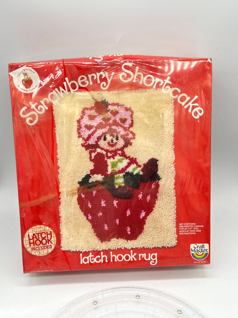 Puede incluir: Un kit de alfombra de gancho de cierre rojo y blanco con Strawberry Shortcake en una cesta de fresas. El kit incluye lienzo preimpreso, hilo acr&iacute;lico e instrucciones. Marca Craft Master.