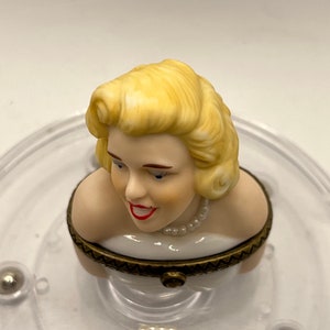 May include: A porcelain figurine of a woman with blonde hair wearing a white dress and a pearl necklace. The figurine is part of a music box.