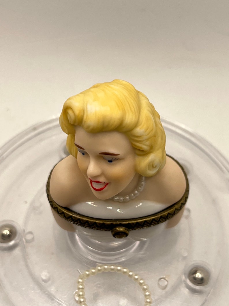 May include: A porcelain figurine of a woman with blonde hair wearing a white dress and a pearl necklace. The figurine is part of a music box.