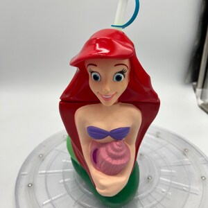 Op de afbeelding: Een rode, groene en paarse plastic beker in de vorm van Ariel, de Kleine Zeemeermin, die een roze schelp vasthoudt. De beker heeft een blauwe en witte rietje.