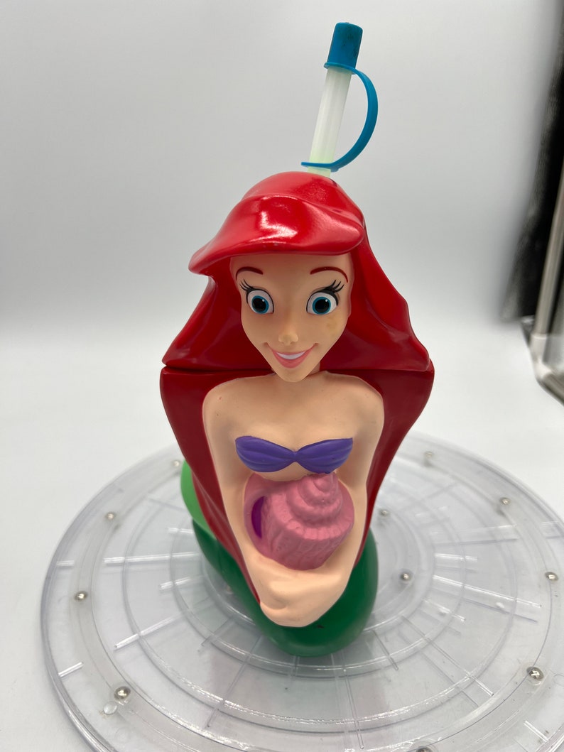 Op de afbeelding: Een rode, groene en paarse plastic beker in de vorm van Ariel, de Kleine Zeemeermin, die een roze schelp vasthoudt. De beker heeft een blauwe en witte rietje.