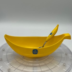 Puede incluir: Un bol amarillo con forma de plátano con una pequeña cuchara en su interior. El bol tiene el logotipo de la banana Chiquita en un lado. El bol está sobre una plataforma giratoria de plástico transparente.