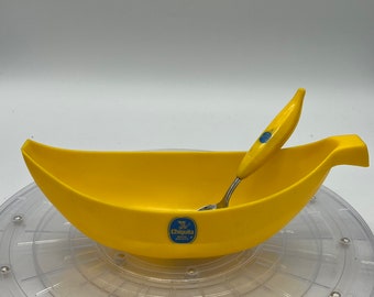 May include: A yellow banana-shaped bowl with a small spoon inside. The bowl has the Chiquita banana logo on the side. The bowl is sitting on a clear plastic turntable.