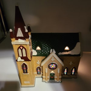 K&ouml;nnte beinhalten: Eine kleine Keramikkirche mit einem braunen Turm, einem gr&uuml;nen Dach und Buntglasfenstern. Die Kirche hat einen schneebedeckten wei&szlig;en Sockel und einen Kranz an der Eingangst&uuml;r.
