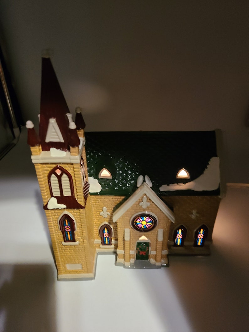K&ouml;nnte beinhalten: Eine kleine Keramikkirche mit einem braunen Turm, einem gr&uuml;nen Dach und Buntglasfenstern. Die Kirche hat einen schneebedeckten wei&szlig;en Sockel und einen Kranz an der Eingangst&uuml;r.