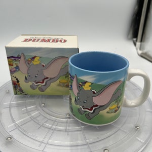 Puede incluir: Una taza de cerámica azul y blanca con una imagen de Dumbo el elefante de Dumbo de Disney. La taza tiene un asa y está sentada sobre una plataforma giratoria de plástico transparente. La caja de la taza está en el fondo y dice "Disney's Dumbo" en ella.