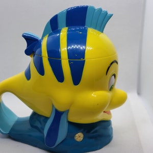 Puede incluir: Una taza de pl&aacute;stico amarilla y azul con forma del personaje Flounder de la pel&iacute;cula de Disney La Sirenita. La taza tiene un asa con forma de la cola de Flounder.