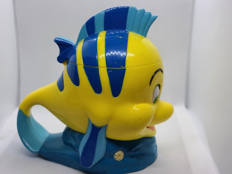 Puede incluir: Una taza de pl&aacute;stico amarilla y azul con forma del personaje Flounder de la pel&iacute;cula de Disney La Sirenita. La taza tiene un asa con forma de la cola de Flounder.