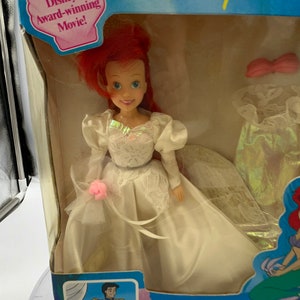 May include: Ariel, the Little Mermaid, is dressed in a white wedding gown with a pink flower. She is holding a bouquet of pink flowers. The doll is packaged in a box with a picture of Ariel and Prince Eric on their wedding day. The box also includes the text "Ariel The Beautiful Bride" and "The Little Mermaid".
