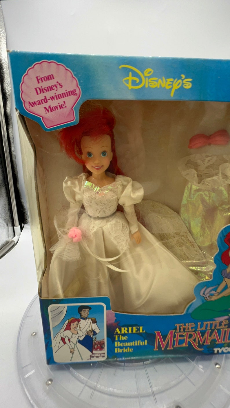 May include: Ariel, the Little Mermaid, is dressed in a white wedding gown with a pink flower. She is holding a bouquet of pink flowers. The doll is packaged in a box with a picture of Ariel and Prince Eric on their wedding day. The box also includes the text "Ariel The Beautiful Bride" and "The Little Mermaid".