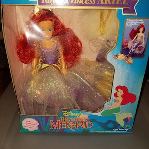 May include: A plastic doll of Ariel from Disney's The Little Mermaid, dressed in a purple and gold gown with a crown. The doll is packaged in a box with a picture of Ariel on the front. The box also includes the text "Disney's The Little Mermaid" and "Ages 3 and up".