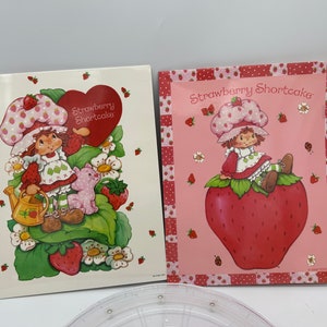 May include: Two vintage Strawberry Shortcake cards. The first card features Strawberry Shortcake holding a heart-shaped sign that says "Strawberry Shortcake" and a small pig. The second card features Strawberry Shortcake sitting on a large strawberry with the text "Strawberry Shortcake".