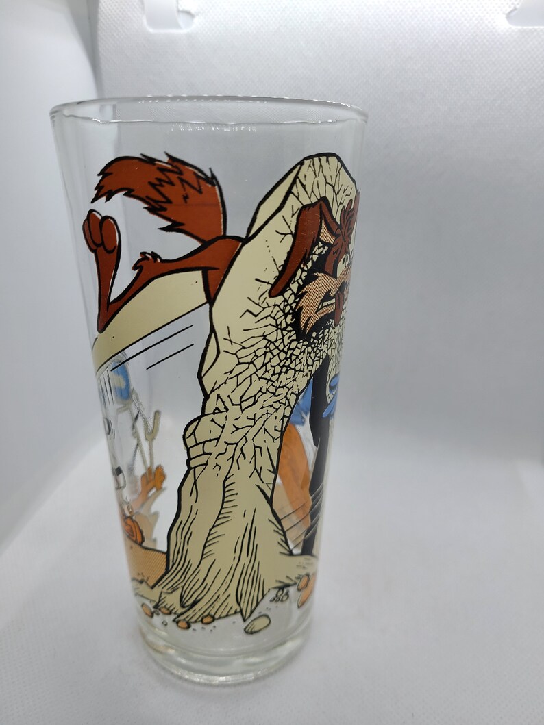May include: A clear glass featuring a cartoon image of Wile E. Coyote and the Road Runner. The Road Runner is running away from Wile E. Coyote, who is holding a large rock. The glass is a vintage collectible.