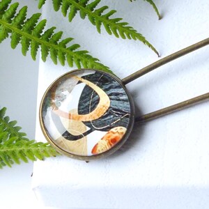 May include: An antique brass safety pin with a decorative, circular head. The head displays an abstract design in black, white, and orange. The pin is set against a white background with green fern leaves.