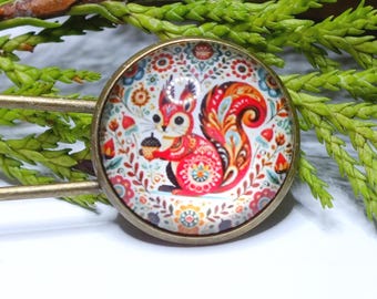 May include: A decorative hair pin with a round, bronze-colored charm featuring a colorful squirrel holding an acorn. The squirrel is red, white, and orange, set against a floral background. Green foliage and a white surface are in the background.