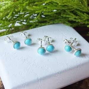 May include: Several pairs of turquoise stud earrings with silver posts are displayed on a white surface. The earrings have a round, light blue stone surrounded by a silver bezel setting. Green foliage is visible in the background.