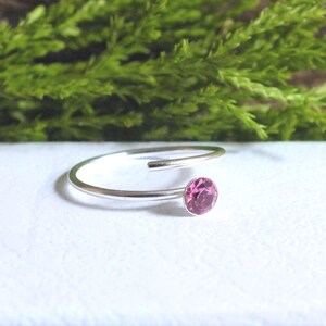 Peut inclure: Une bague argentée ornée d'une pierre rose. La bague présente un design minimaliste, avec une seule pierre rose ronde sertie sur un fin anneau. La bague est présentée sur une surface blanche avec un feuillage vert en arrière-plan.