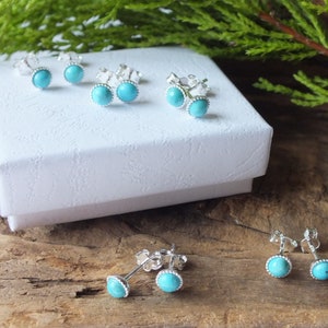 May include: Several pairs of turquoise stud earrings with silver settings are displayed on a white box and a wooden surface. The earrings feature a round, light blue stone surrounded by a beaded silver border. The earrings are a classic jewelry item.