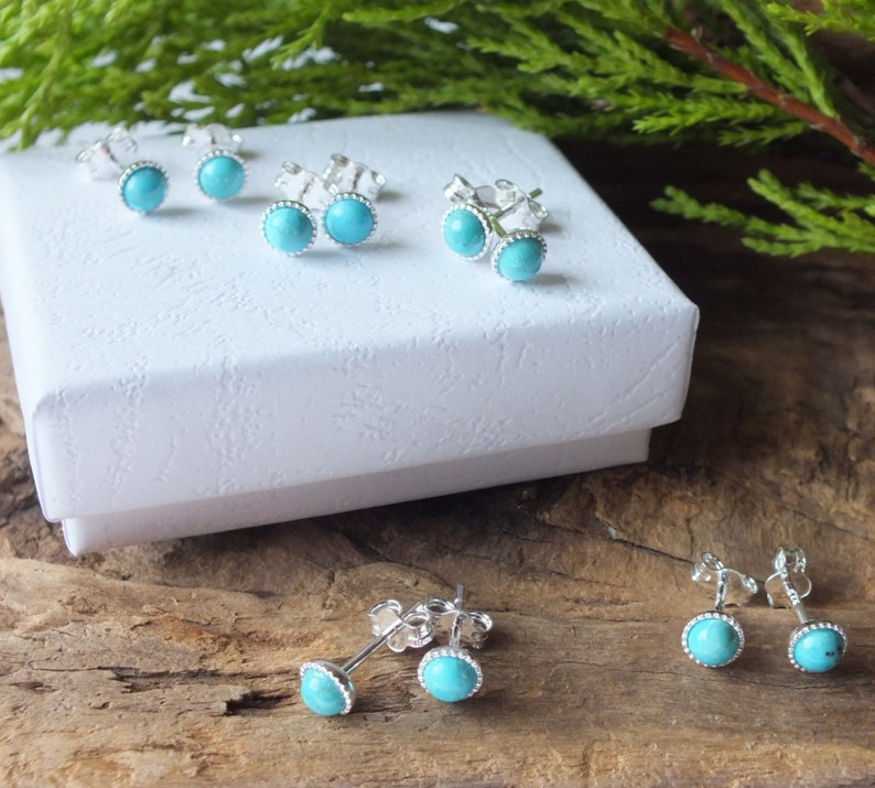 May include: Several pairs of turquoise stud earrings with silver settings are displayed on a white box and a wooden surface. The earrings feature a round, light blue stone surrounded by a beaded silver border. The earrings are a classic jewelry item.