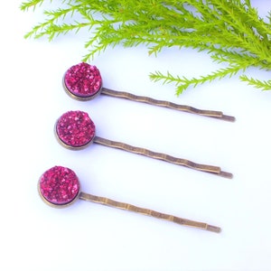Può includere: Tre forcine per capelli in bronzo antico, ciascuna con una pietra druzy rotonda color magenta. Le forcine sono disposte su una superficie bianca con fogliame verde sullo sfondo. Le forcine hanno un design strutturato e ondulato.