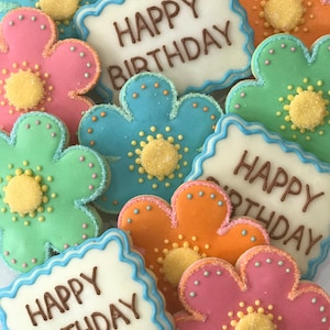 May include: Assortment of decorated sugar cookies with a flower design. The cookies are decorated with blue, green, pink, and orange frosting. The cookies have the words "Happy Birthday" written on them.