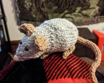 May include: A gray and brown knitted mouse with a long tail. The mouse is sitting on a red and black plaid blanket.