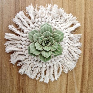 Peut inclure: Un dessous de verre en macramé fait à la main avec un motif de succulente verte au centre. Le dessous de verre est rond, avec un bord frangé de couleur crème et une succulente en céramique détaillée au centre. Posé sur une surface en bois.