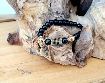 Puede incluir: Pulsera de cuentas negras con un dije dorado con un diseño cuadrado negro.