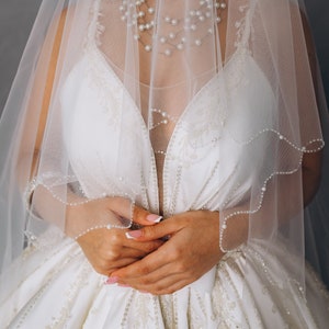 May include: A bride wearing a white wedding dress with a veil and a pearl necklace. The dress has a beaded design and the veil is trimmed with pearls.