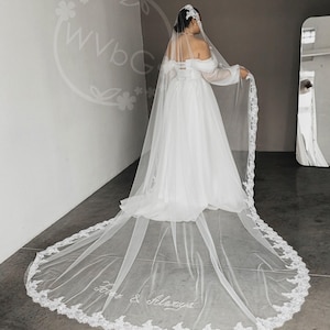 Pode incluir: Vestido de noiva branco com uma longa cauda e véu. O véu tem uma delicada borda de renda e as palavras "Forever & Always" bordadas. A noiva usa um adorno de cabeça e o vestido tem mangas compridas.