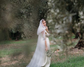 May include: A bride in a white wedding dress with a long veil, holding a bouquet of greenery. The dress has a flowing skirt and a fitted bodice. The veil is draped over the head and shoulders. The background is a green field with trees.