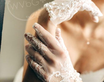 Könnte beinhalten: Zarte weiße Spitzenhandschuhe mit floralen Mustern und Perlen am Handgelenk. Die Handschuhe sind aus einem transparenten, offen gewebten Stoff gefertigt, ideal für Hochzeiten oder formelle Anlässe.