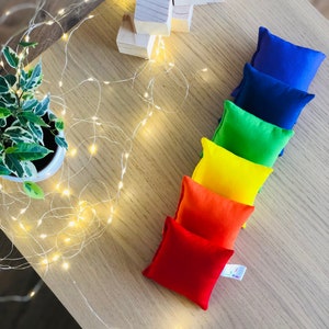 Puede incluir: Un conjunto de siete bolsas de frijoles coloridas dispuestas en un patr&oacute;n de arco&iacute;ris sobre una superficie de madera. Las bolsas de frijoles est&aacute;n hechas de tela y est&aacute;n llenas de frijoles. Tambi&eacute;n hay bloques de madera en el fondo.