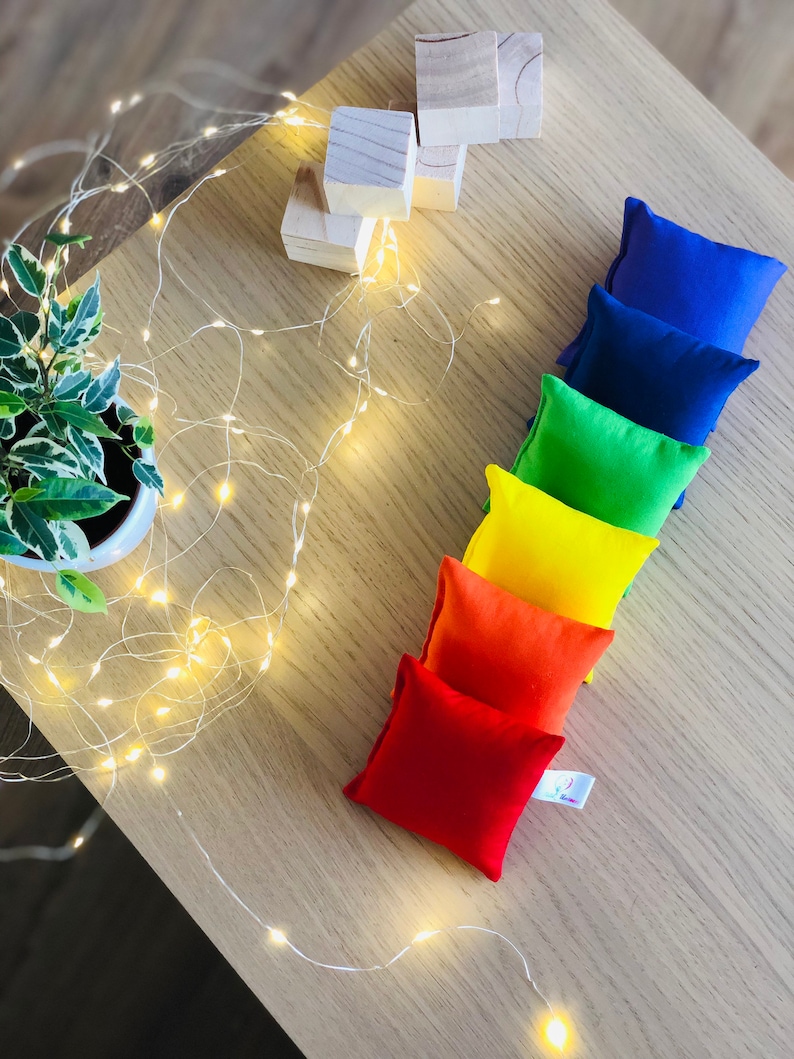 Puede incluir: Un conjunto de siete bolsas de frijoles coloridas dispuestas en un patr&oacute;n de arco&iacute;ris sobre una superficie de madera. Las bolsas de frijoles est&aacute;n hechas de tela y est&aacute;n llenas de frijoles. Tambi&eacute;n hay bloques de madera en el fondo.