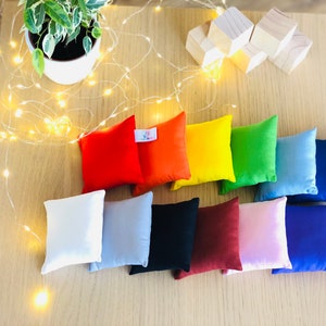 Puede incluir: Diez peque&ntilde;os cojines cuadrados de varios colores, incluyendo rojo, naranja, amarillo, verde, azul, blanco, gris, negro, marr&oacute;n y rosa. Los cojines est&aacute;n dispuestos en una fila sobre una superficie de madera.