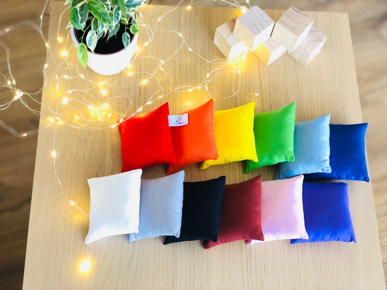 Puede incluir: Diez peque&ntilde;os cojines cuadrados de varios colores, incluyendo rojo, naranja, amarillo, verde, azul, blanco, gris, negro, marr&oacute;n y rosa. Los cojines est&aacute;n dispuestos en una fila sobre una superficie de madera.