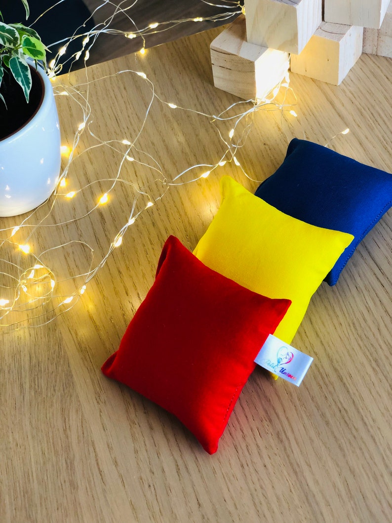 Puede incluir: Tres bolsas de frijoles de tela en rojo, amarillo y azul. Las bolsas de frijoles est&aacute;n sobre una superficie de madera con una cadena de luces blancas. Las bolsas de frijoles tienen una etiqueta blanca con un coraz&oacute;n rojo y el texto "Play Time!"