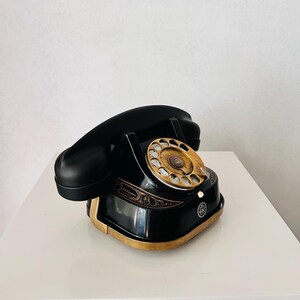 May include: A vintage black rotary phone with a gold-colored dial and base trim. The phone features a curved handset and decorative detailing. The phone is a classic design, likely from the mid-20th century.