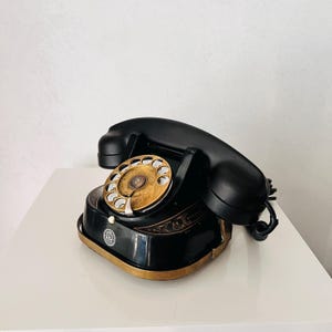 May include: A vintage black rotary phone with a gold dial and accents. The phone has a classic design with a handset and a base with decorative detailing. The phone is sitting on a white surface.