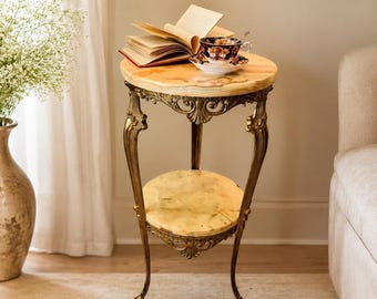 May include: A two-tiered round side table featuring a polished light tan marble top and a matching lower shelf. The table is supported by three ornate, curved brass-toned metal legs with intricate scrollwork details at the top and base.
