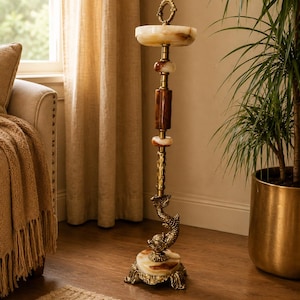 May include: A tall floor-standing ashtray featuring a circular cream-colored stone tray at the top, a central column with brown and cream stone accents, and a brass base decorated with a sculpted dolphin figure and ornate metal feet.