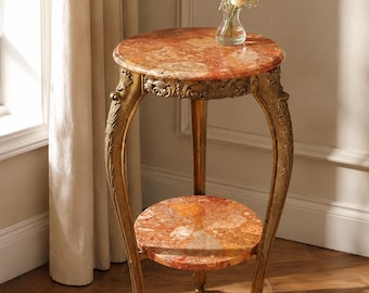 May include: A round side table featuring two tiers of polished, multi-colored orange and red marble surfaces. The table is supported by three curved, gold-toned metal legs with intricate embossed detailing along the upper frame and feet.