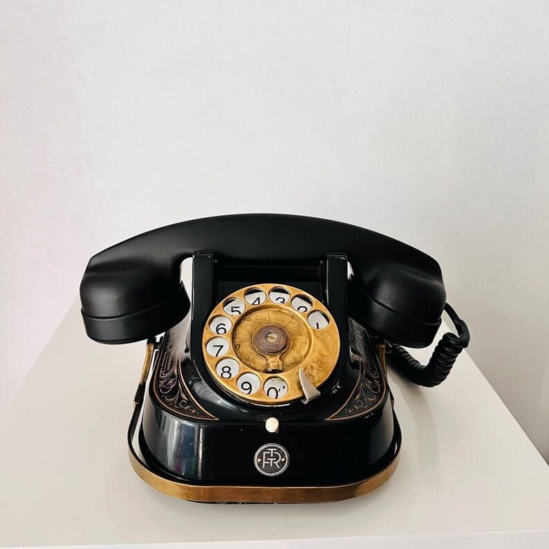 May include: A vintage black rotary dial telephone with a gold-colored dial and accents. The handset rests on the base, and a coiled cord connects it to the phone. The phone has an ornate design.