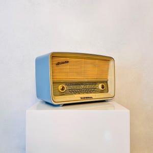 May include: A vintage radio with a light blue casing and a tan speaker grill. The radio has gold-colored knobs and trim, and the word "Astoria" is printed on the top. The radio is displayed on a white pedestal.