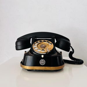 May include: A vintage black rotary phone with a gold dial and trim. The handset rests on the cradle, and a coiled cord extends to the right. The phone has a classic design, with a circular dial and a base with decorative detailing.