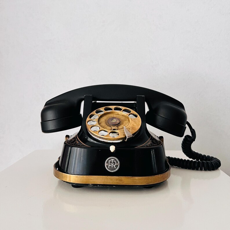 May include: A vintage black rotary phone with a gold dial and trim. The handset rests on the cradle, and a coiled cord extends to the right. The phone has a classic design, with a circular dial and a base with decorative detailing.
